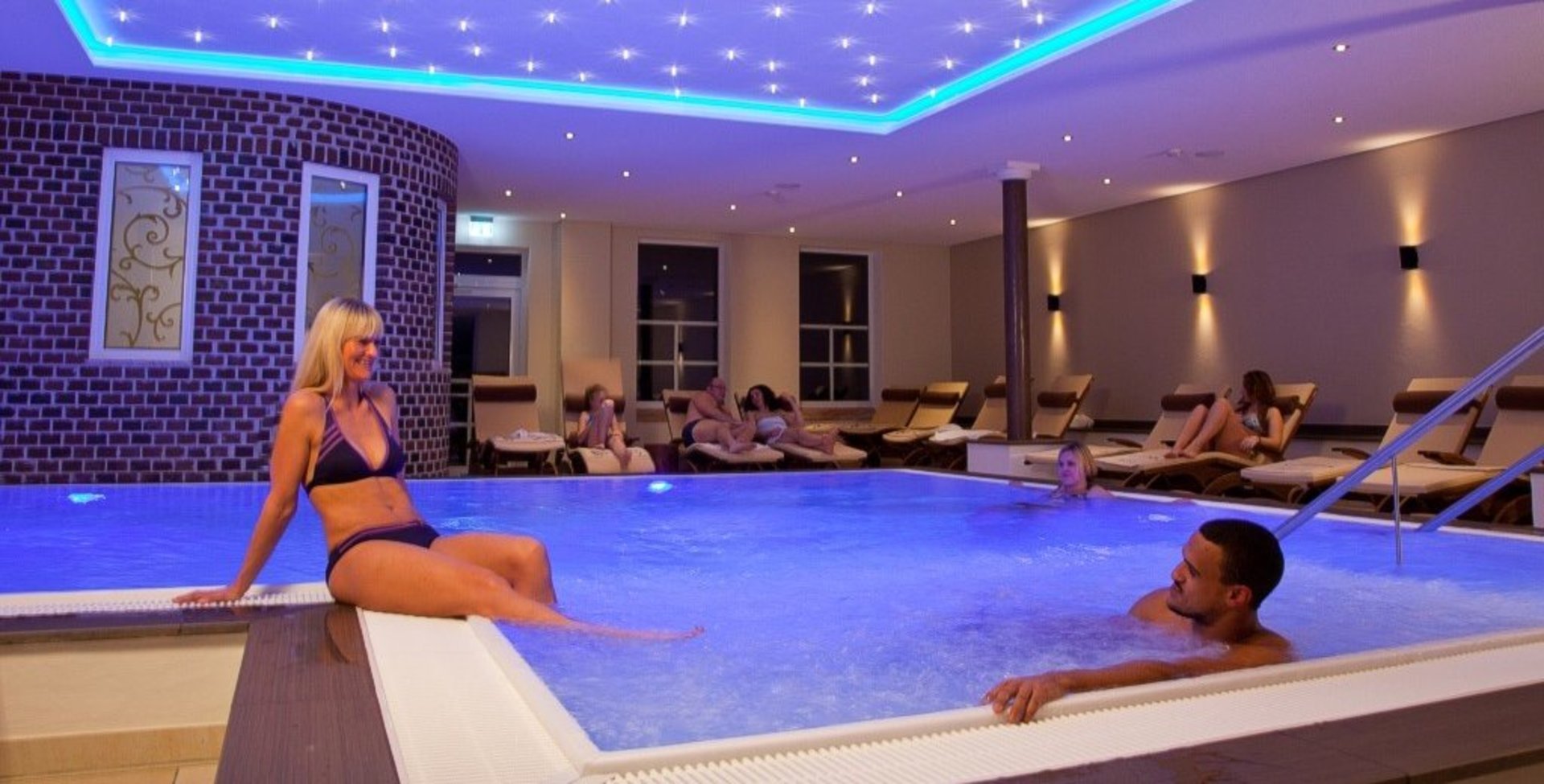 Wellness-Hotels Blick auf den Indoorpool. Drei Gäste sind im Wasser, auf den Liegen dahinter ruhen sich einige Leute aus.
