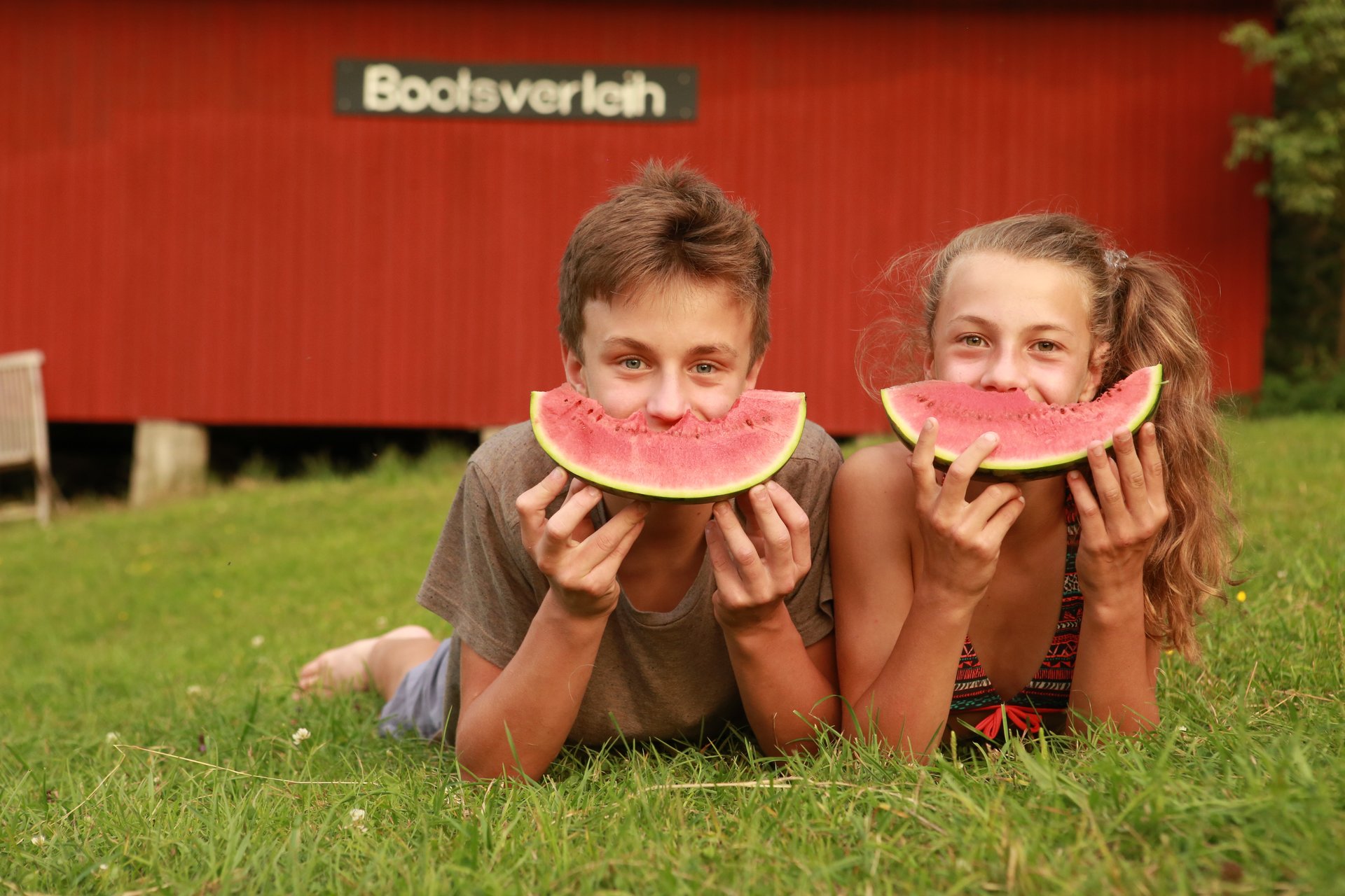 Kurbeitrag zwei Kinder liegen in der Wiese und halten sich eine Melonenscheibe vor den Mund, so dass es aussieht als hätten sie ein breites Grinsen im Gesicht. Im Hintergrund sieht man die Scheune in der die Boote für den Bootsverleih gelagert werden.