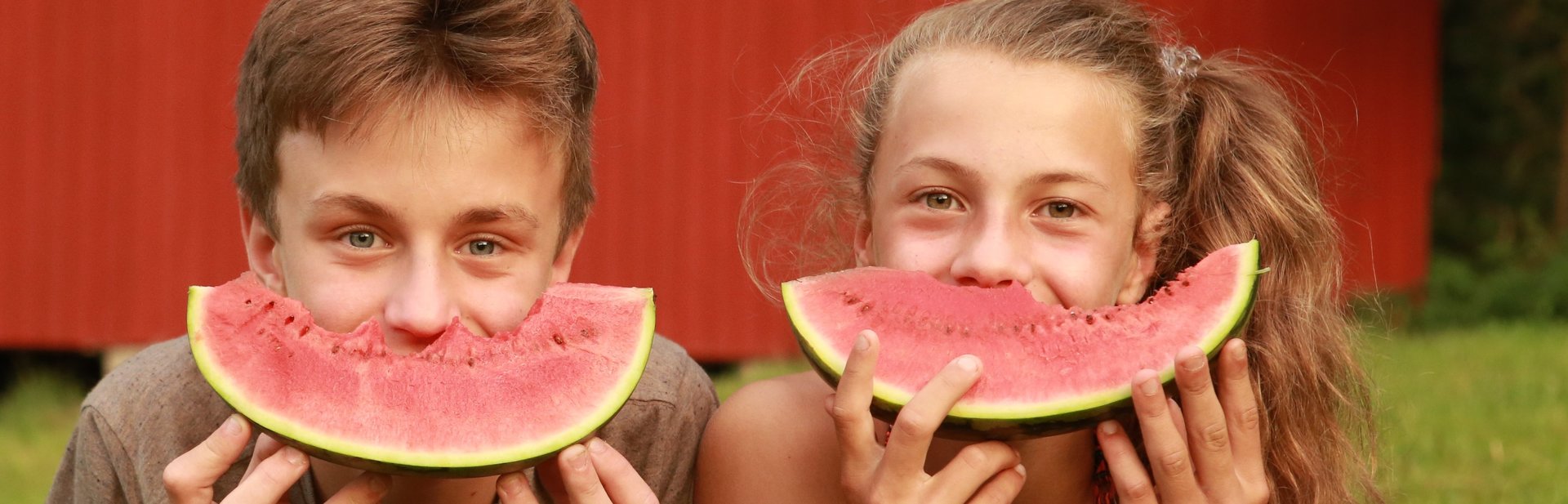 Kurbeitrag zwei Kinder liegen in der Wiese und halten sich eine Melonenscheibe vor den Mund, so dass es aussieht als hätten sie ein breites Grinsen im Gesicht. Im Hintergrund sieht man die Scheune in der die Boote für den Bootsverleih gelagert werden.