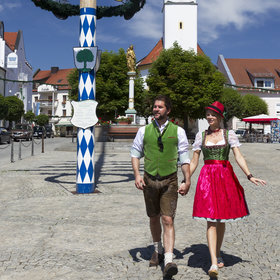 Paar in Tracht marschiert über den Stadtplatz. Im Hintergrund sieht man einen Maibaum und eine Kirche