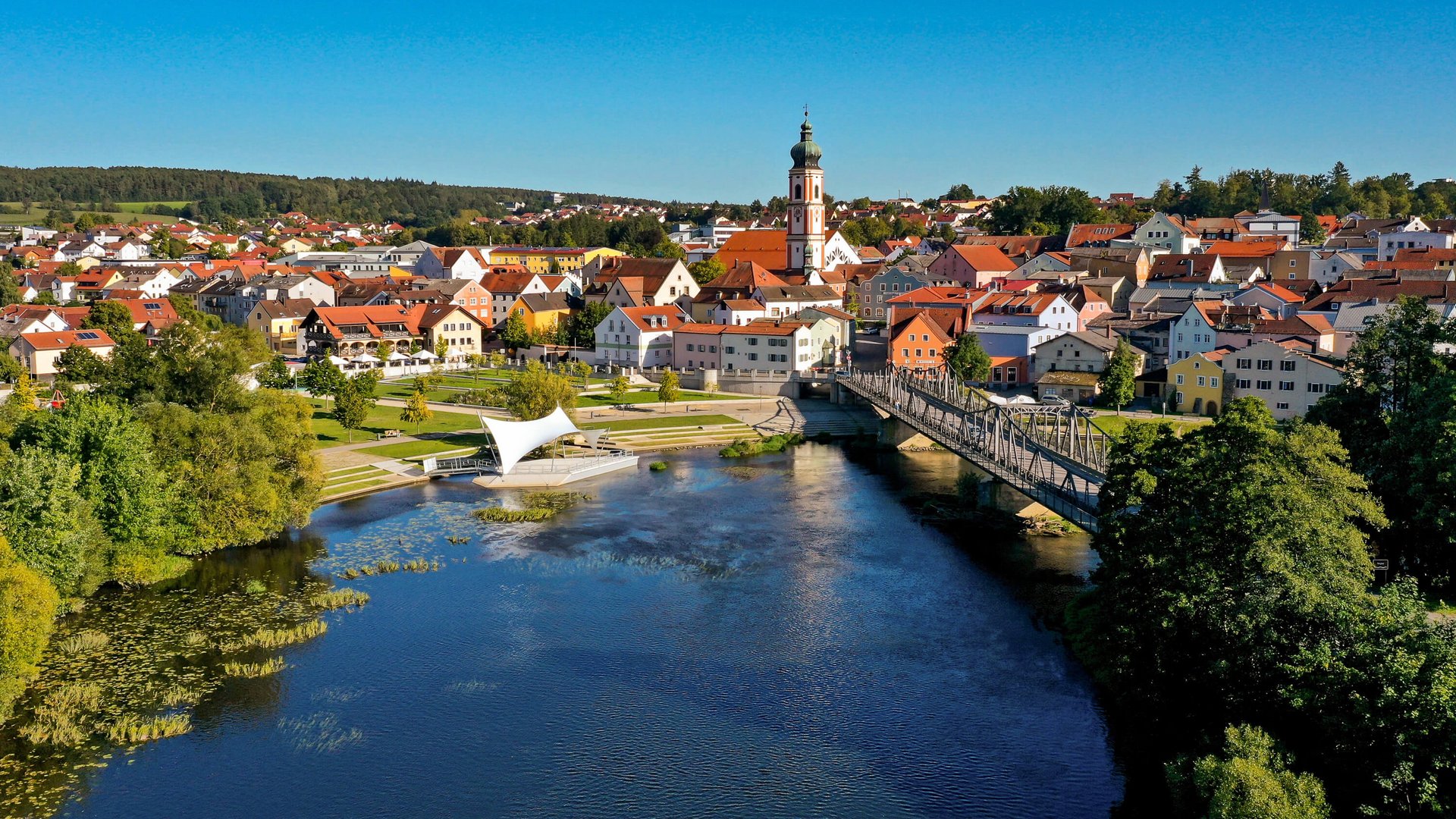 Drohnenaufnahme über den Fluss Regen mit Regenpromenade, angrenzender Rodinger Altstadt, Kirchturm und Flussbühne Roding.