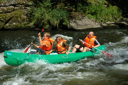 Blick auf ein grünes Kanu das auf dem Fluss Regen entlang fährt darin sitzt ein Vater mit seinen zwei Kindern im Kanu. Die drei haben orange Schwimmwesten an. Vater und Sohn rudern