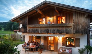 Bayerisches Haus mit gemütlicher Atmosphäre: Personen sitzen an einem Holztisch im Freien, während Kinder vom Holzbalkon hinunterschauen.