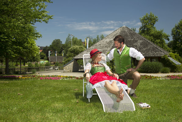 Paar in Tracht sitzt auf einer Liege in der Wiese im Kurpark. Im Hintergrund sieht man den Pavillon und ein buntes Blumenbeet