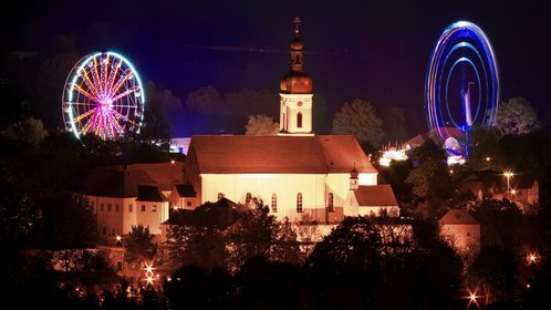 Abendlicher Blick auf beleuchtete Kirchenburg von Bad Kötzting, umrahmt von bunt beleuchteten Fahrgeschäften