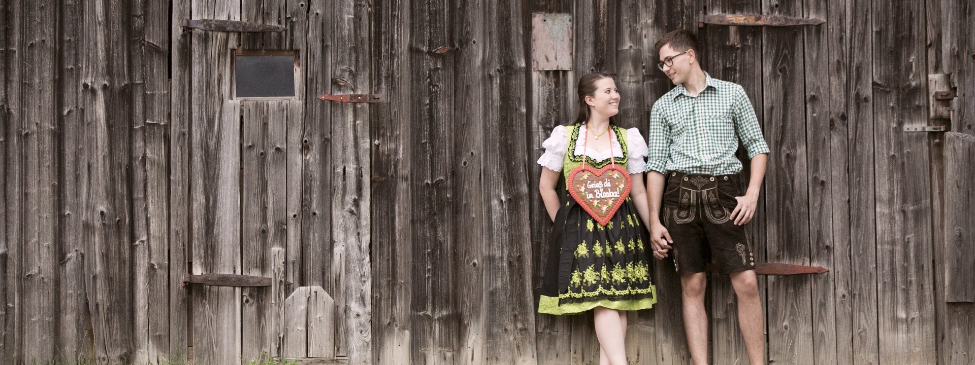 Griaß Paar in Tracht vor einer Holzwand mit einem Lebkuchenherz mit der Aufschrift "Griaß di in Bloaba"