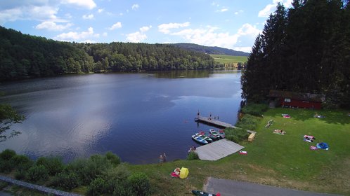 Luftaufnahme vom See und der Badestelle, auf der Liegewiese und dem Steg befinden sich Badegäste im Wasser schwimmen fünf bunte Boote
