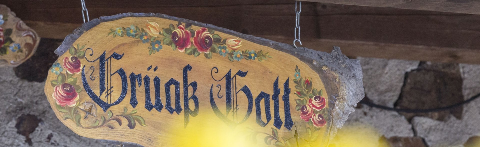 Foto von einem verziertem Holzschild, auf dem "Grüß Gott" steht, im Vordergrund sieht man verschwommen gelbe Blumen