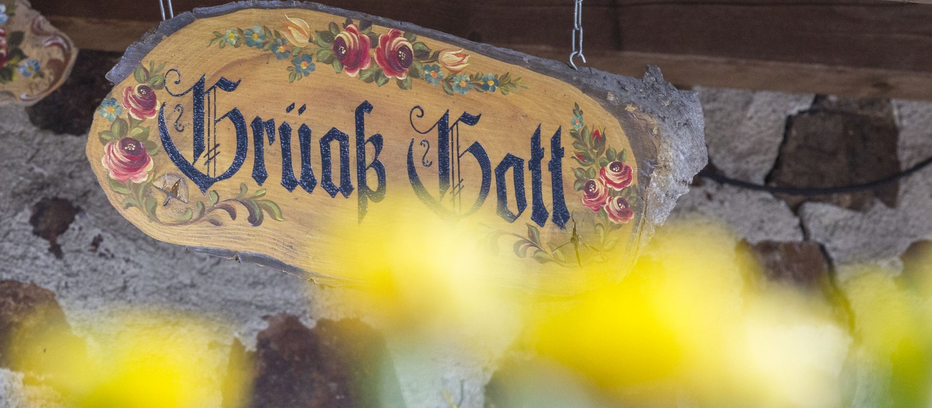 Foto von einem verziertem Holzschild, auf dem "Grüß Gott" steht, im Vordergrund sieht man verschwommen gelbe Blumen