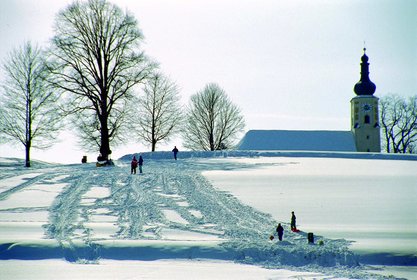 Blick auf die verschneite Kirche in Weißenregen, den Berg fahren Kinder mit dem Schlitten herunter